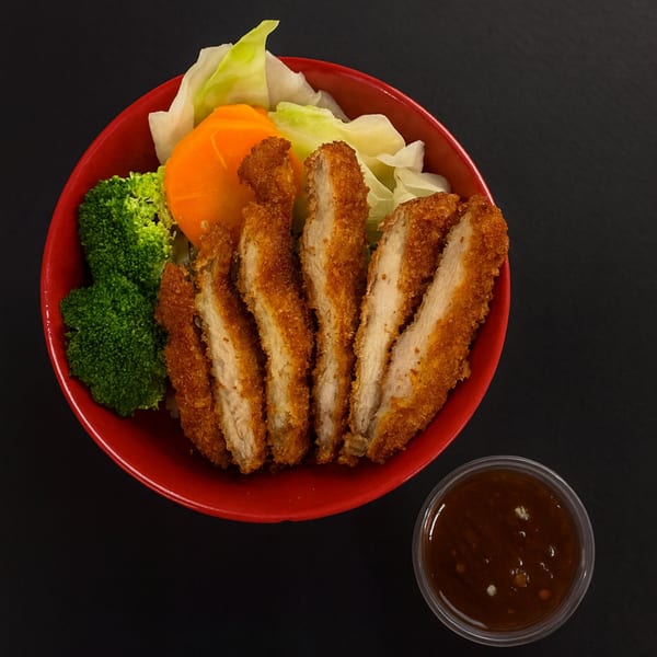 Crispy chicken katsu slices with broccoli, cabbage, and carrots in a red bowl, with a side of sauce.