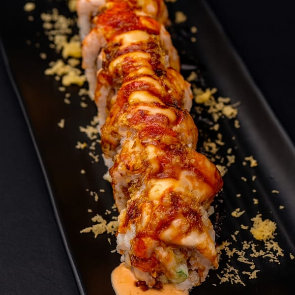 A close-up of a sushi roll with creamy sauce and crunchy toppings on a black plate.