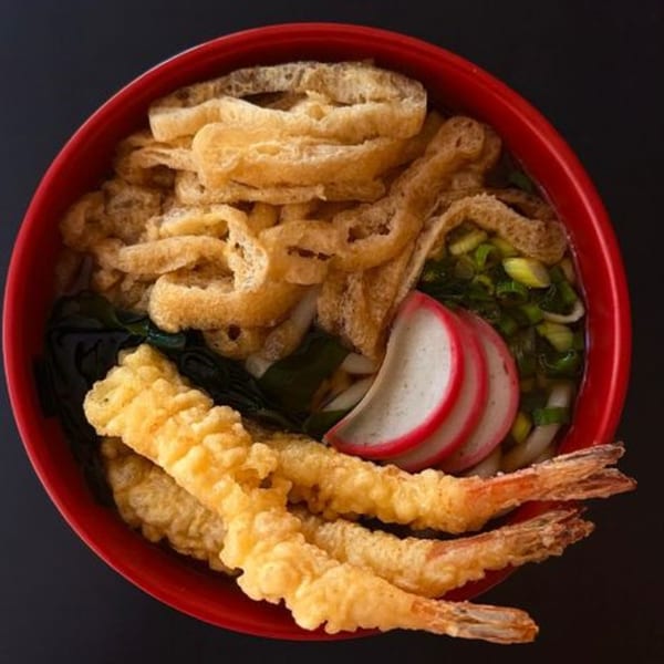 A red bowl of Japanese udon noodles with tempura shrimp, fish cake, and green onions.
