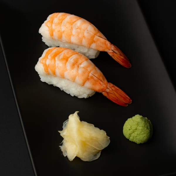Two shrimp nigiri sushi pieces with pickled ginger and wasabi on a black plate.