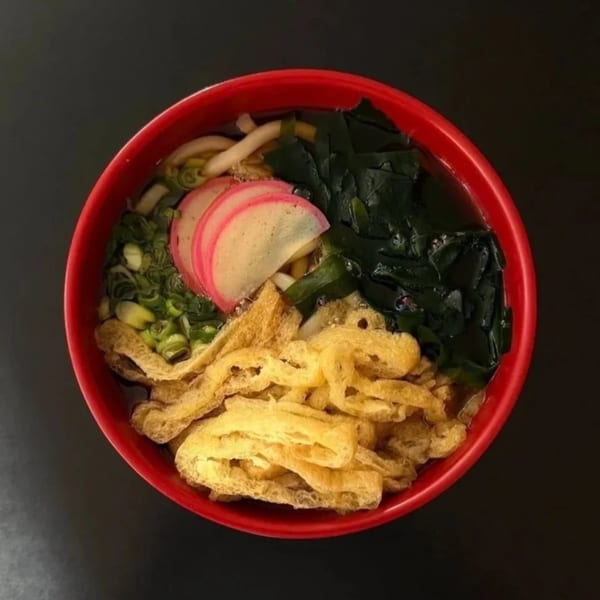 A red bowl of Japanese udon noodles with fish cake, seaweed, green onions, and fried tofu.